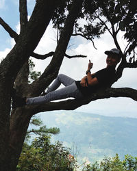 Young man sitting by tree against sky