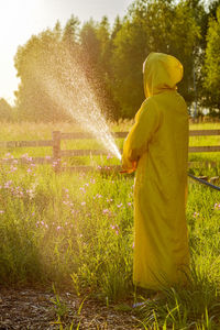 A man in a yellow raincoat spraying a green lawn from a hose spraying water