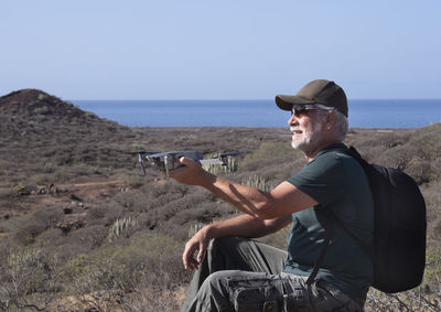 Smiling man wearing cap holding drone sitting on land against sky