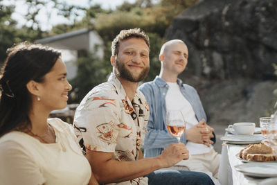 Portrait of smiling young man holding wineglass while sitting with friends during dinner party at cafe