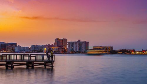 Illuminated buildings by river against sky at sunset