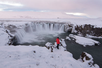 Rear view of woman standing on frozen cliff by waterfall against cloudy sky