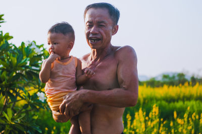 Shirtless man carrying son while standing against sky