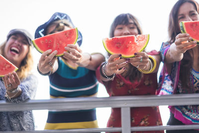 Low angle view of smiling mid adult friends standing with watermelon slices at railing against sky