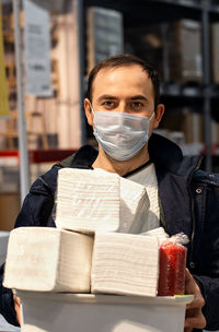 Man wearing medical face mask is holding many packs of tissues or paper towels 