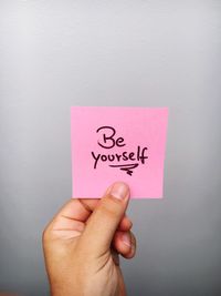 Cropped hand of person holding be yourself text on adhesive note against gray background