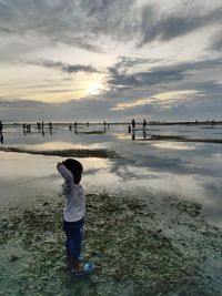 Rear view of boy standing on beach against sky during sunset