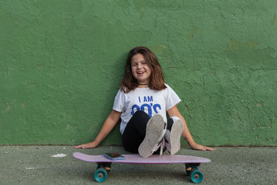 Portrait of girl sitting on skateboard against wall