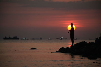 Silhouette man standing on rock by sea against sky during sunset