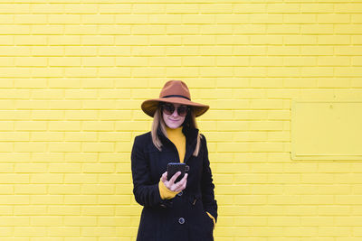 Fashionable mid adult woman using mobile phone while standing against yellow wall