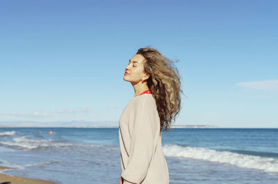 Young woman with curly hair standing at beach against clear sky