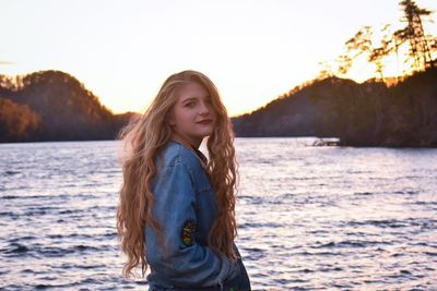 Portrait of smiling young woman standing by lake against sky during sunset