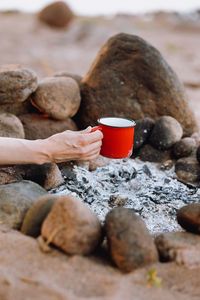 A woman's hand holds a red cup of tea or coffee on the background of  a burning campfire on a hike