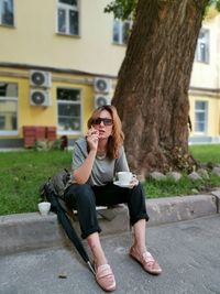 Full length of woman holding coffee cup while sitting on footpath