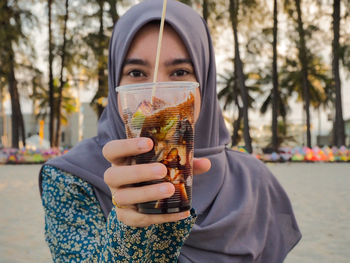 Portrait of young woman holding drink while standing at beach during sunset