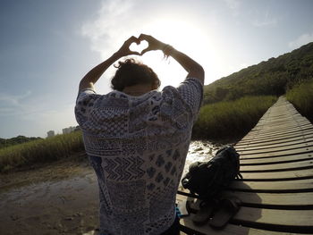 Rear view of man making heart shape on boardwalk against sky