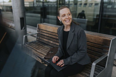 Happy businesswoman sitting with laptop on bench while waiting at railroad station