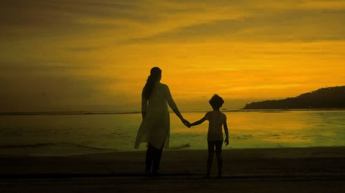 Silhouette friends standing on beach against orange sky