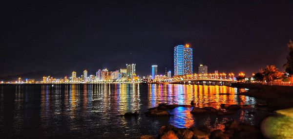 Illuminated buildings by sea against sky at night