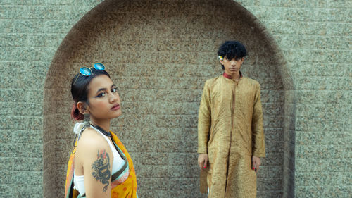 A boy and a girl stand infront of an arch shaped wall, in traditional wear.