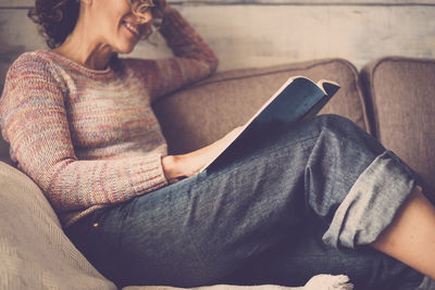 Midsection of young woman reading book while relaxing on sofa at home