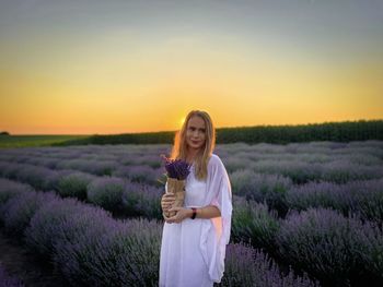 Young woman in white dress standing on a field of lavender at sunset and holding a lavender bouquet