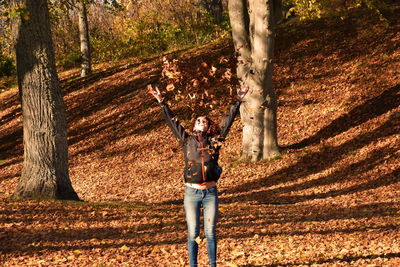 Smiling woman throwing dry leaves while standing against tree trunk in forest