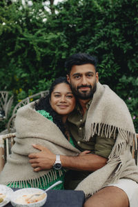 Portrait of man and woman covered in blanket embracing each other while sitting on bench
