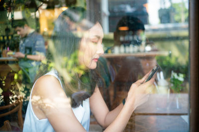 Side view of woman using mobile phone while sitting at table seen through glass window