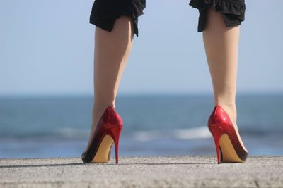 Low section of woman wearing red high heels standing against sea