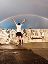 Rear view of man with arms raised standing on road against graffiti wall