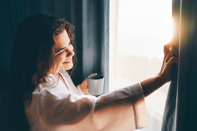 Curly haired lady in white bathrobe opens curtains to look outside large window drinking hot coffee