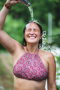 Happy female standing with closed eyes in top and pouring water from bottle on head against green trees in park in summer time