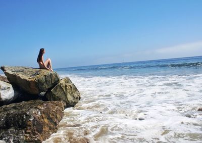 Side view of young woman sitting on rock by sea against sky
