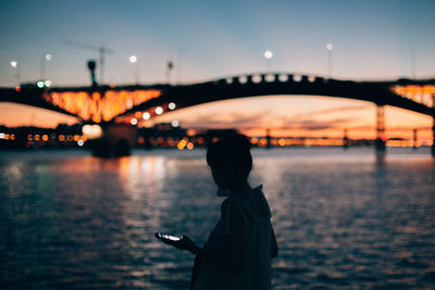 Side view of woman with smart phone walking by river in city during sunset