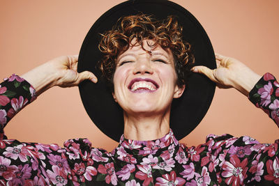 Optimistic young beautiful female model with curly hair wearing stylish black hat and colorful blouse with closed eyes smiling while standing against orange background