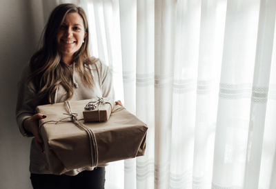 Portrait of smiling young woman holding gift box standing by window at home