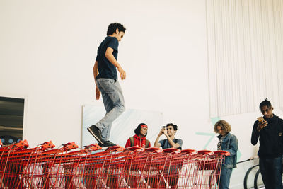 Young man photographing friend walking on stacked shopping carts outside mall