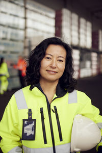 Portrait of confident female worker in reflective clothing holding hardhat in factory