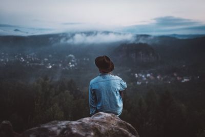 Rear view of man sitting on rock against sky at dusk