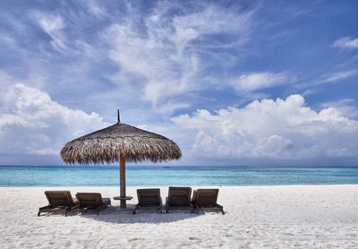 Lounge chairs under thatched roof at beach against sky