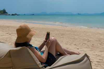 Young woman sitting on bean bag on beach and using smartphone to take photo during summer vacation