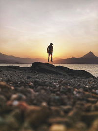 Man standing on rocks at beach against sky during sunset