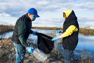 Side view of man and woman picking garbage near lake