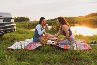 Friends sitting on grass against sky during sunset