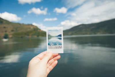 Cropped hand holding photograph by lake against mountains