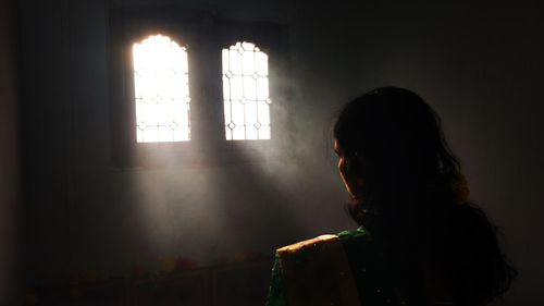 Rear view of woman praying against window in room