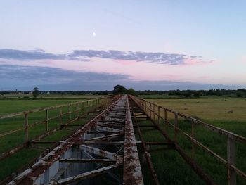 Rusty metal bridge over grassy field against sky at dusk