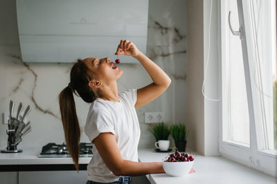 Attractive young woman eats fresh cherries, healthy berries sitting at home in the kitchen