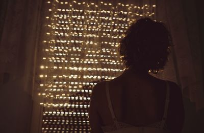 Rear view of woman standing against illuminated window